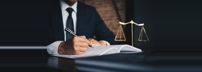 Homme en costume cravate assis à son bureau et corrigeant un document, une balance figurant la justice ou le droit est posée sur son bureau
