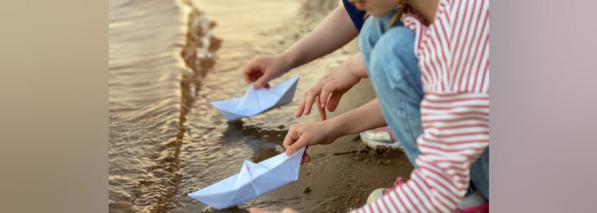 Childrens hands play with paper boats on sea or river in summer at sunset