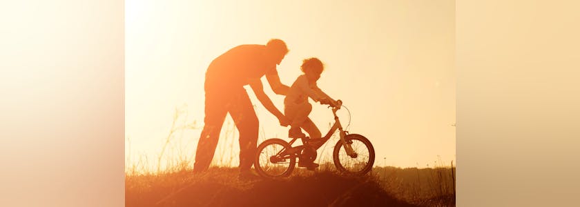 Silhouette of father teaching his daughter to ride a bike at sunset