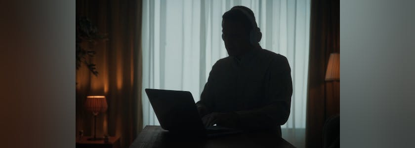 Dark silhouette of a man in wireless headphones at a table in front of a laptop close up. A man works on a laptop, listens to music.